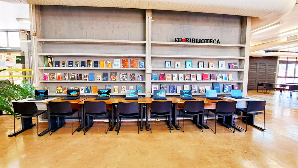 Biblioteca da Unesp: 6 mesas e cadeiras, com 6 notebooks. Estante ao fundo com livros e a frase "eu amo biblioteca".