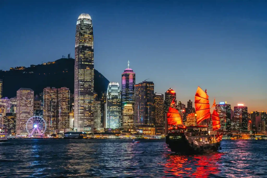 Red-sailed junk boat crossing Victoria Harbour in front of the Hong Kong skyline at dusk.