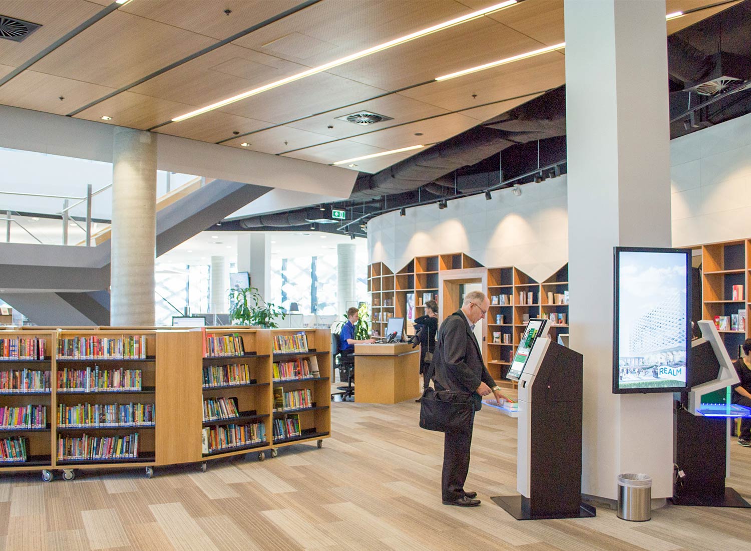 Webinar: Classics Revisited – A Theory of Justice by John Rawls 9 A user interacting with a self-checkout kiosk at Realm Library, Australia, showcasing a modern library layout with bookshelves and digital displays.