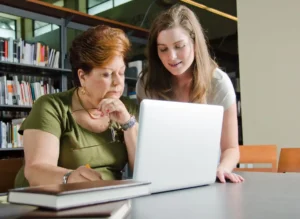 Library staff member helping a patron with a laptop at a study table, with bookshelves in the background
