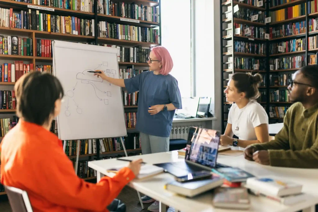 Four library staff members in a meeting, one drawing a diagram on a flip chart, with book stacks behind them.