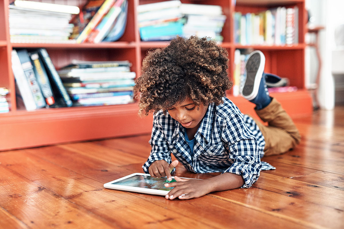 Menino lendo ebook em um tablet deitado de barriga para baixo no chão.